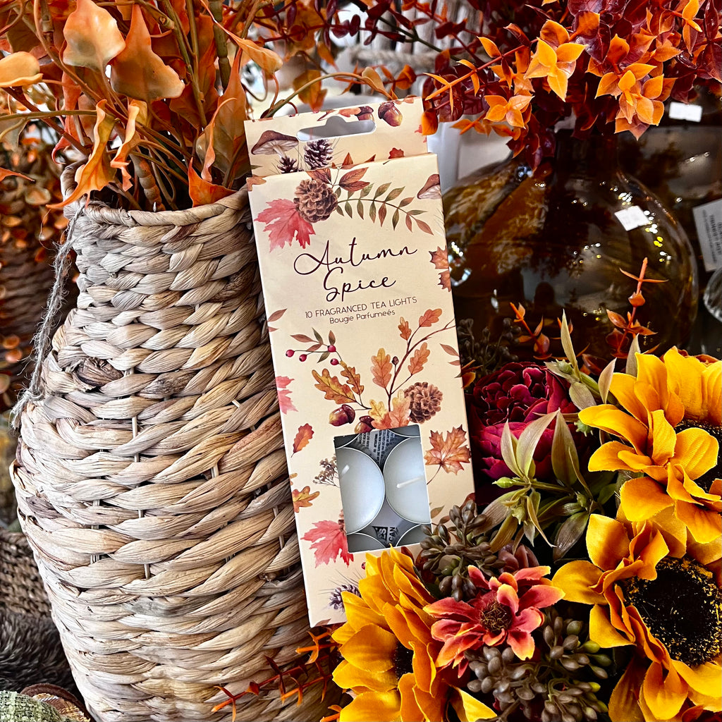 Pack of 'Autumn Spice' scented tea lights with a decorative label, surrounded by autumn-themed flowers and a woven basket.