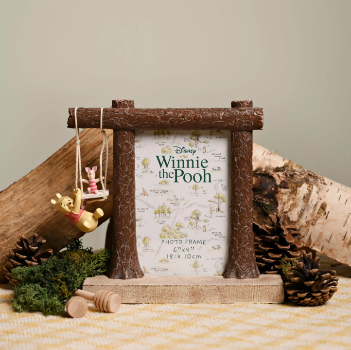 Resin photo frame with fake logs for the frame border on the ides and top. On the top the log extends out to the left where Piglet is on a swing and Winnie the Pooh is hanging from the swing. Frame comes on a wooden style base.