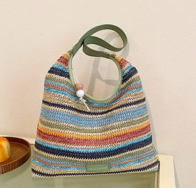 Woven style bag with layers of colours sporadically alternating between shades of blue, a few layers of green, a few layers of red and orange, and neutral beige colours. Green khaki coloured leather shoulder strap.