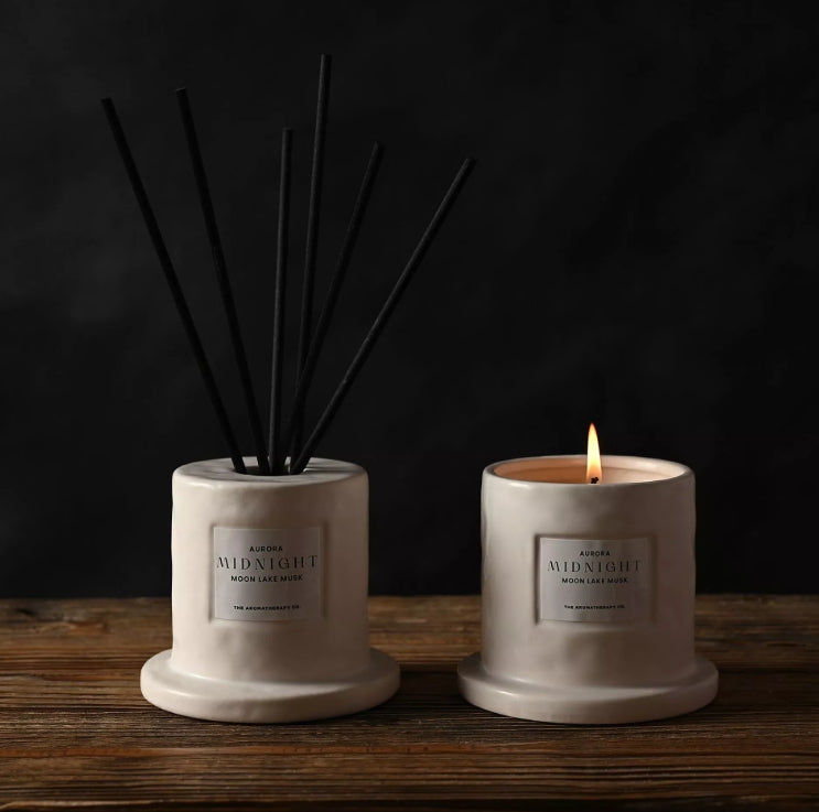 Two diffusers, one with reeds and one with a lit candle, on a wooden surface with a dark background.